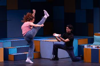 A photo from a performance of "Company," depicting an actress about to kick through a wooden board held by her costar.