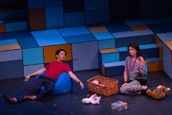 A photo from a performance of "Company," depicting an actor and actress sat on the floor around a bin of spilled toys. The actor holds a cigarette in his hand as the actress speaks.