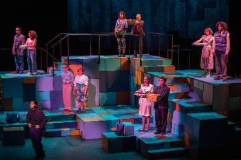 A photo from a performance of "Company," depicting a group of eleven actors standing in couples with one actor stood alone. The couples hold birthday presents and cake towards the actor that stands alone.