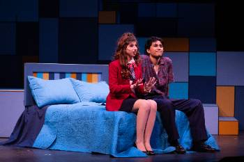 A photo from a performance of "Company," depicting an actor and actress sitting on a blue bed. The actress is mid dialogue as the man listens with a smile.
