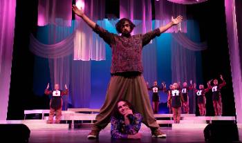 A photo depicting a performance of "Kingdom of Cards," depicting an actor striking a wide, standing pose as their costar smiles happily from his laying position on the floor between their legs.