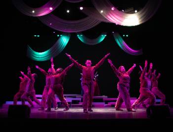 A photo depicting a performance of "Kingdom of Cards," depicting a group of actors posing in a half circle with their arms posed in an upwards "V" shape under harsh pink lighting.