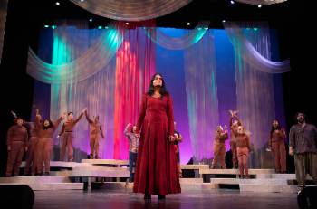 A photo depicting a performance of "Kingdom of Cards," depicting an actress in a red, regal dress standing at a center of a celebrating group.