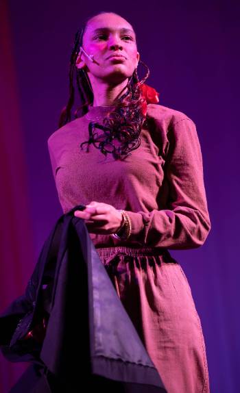 A photo depicting a performance of "Kingdom of Cards," depicting an actress posed proudly with a red ribbon her hair under stark red and blue lighting.