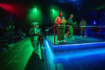A photo from a performance of "Wolf Play," depicting an actor puppeteering a child doll as they walk on the side of a boxing ring. Within the ring, four actors speak to the crowd.