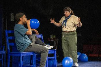 A photo from a performance of "Wolf Play," depicting an actor looking in disappointment at an actor blowing up a blue balloon.