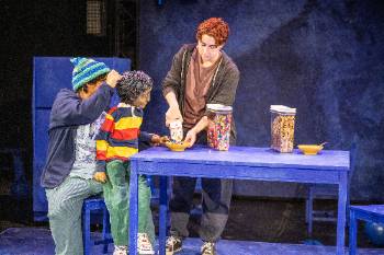 A photo from a performance of "Wolf Play," depicting an actor pouring milk into a bowl of cereal for an actor puppeteering a child doll.