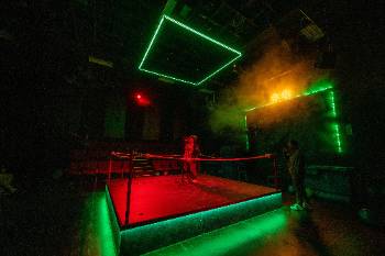 A photo from a performance of "Wolf Play," depicting an actor performing into a standing microphone at the center of a boxing ring, under neon red and green lights.