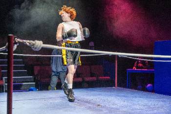 A photo from a performance of "Wolf Play," depicting an actor in a boxing uniform and boxing gloves, posed as though they have just been punched in the side of the face. In the background, another actor watches.