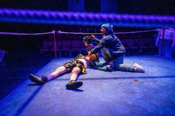 A photo from a performance of "Wolf Play," depicting an actor puppeteering a child doll, kneeling over an actor in a boxing uniform laid on the ground.