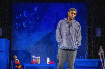 A photo from a performance of "Wolf Play," depicting an actor standing solemnly and looking at the ground. In the background are balloons spelling "Wolf Play" and a child doll sat at a table.