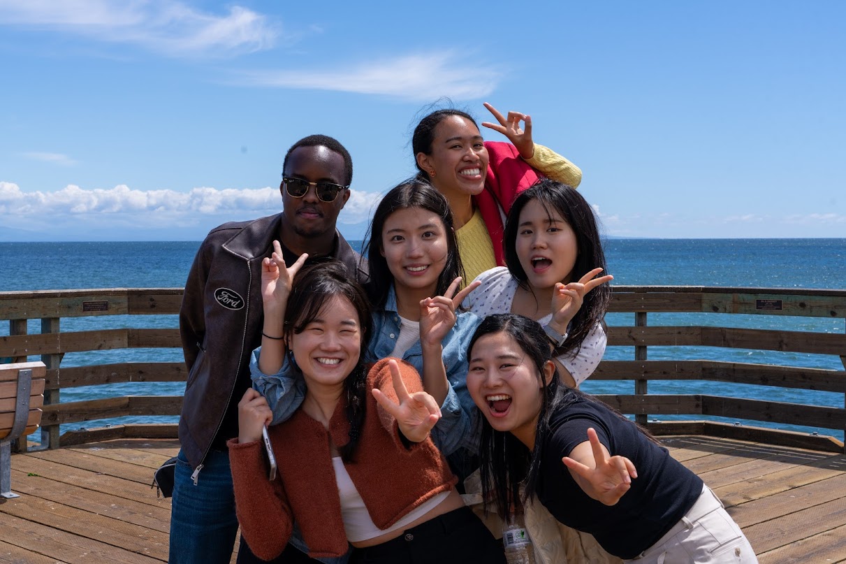 A cohort of students strike joyful poses on a wooden pier overlooking the ocean
