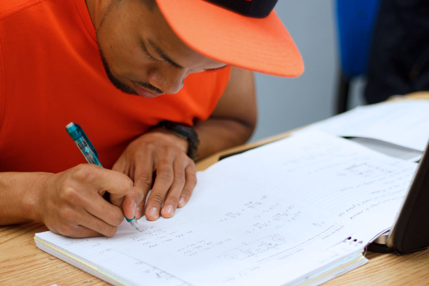 A student is working hard writing in a notebook with a pencil that is filled with difficult mathematics equations.
