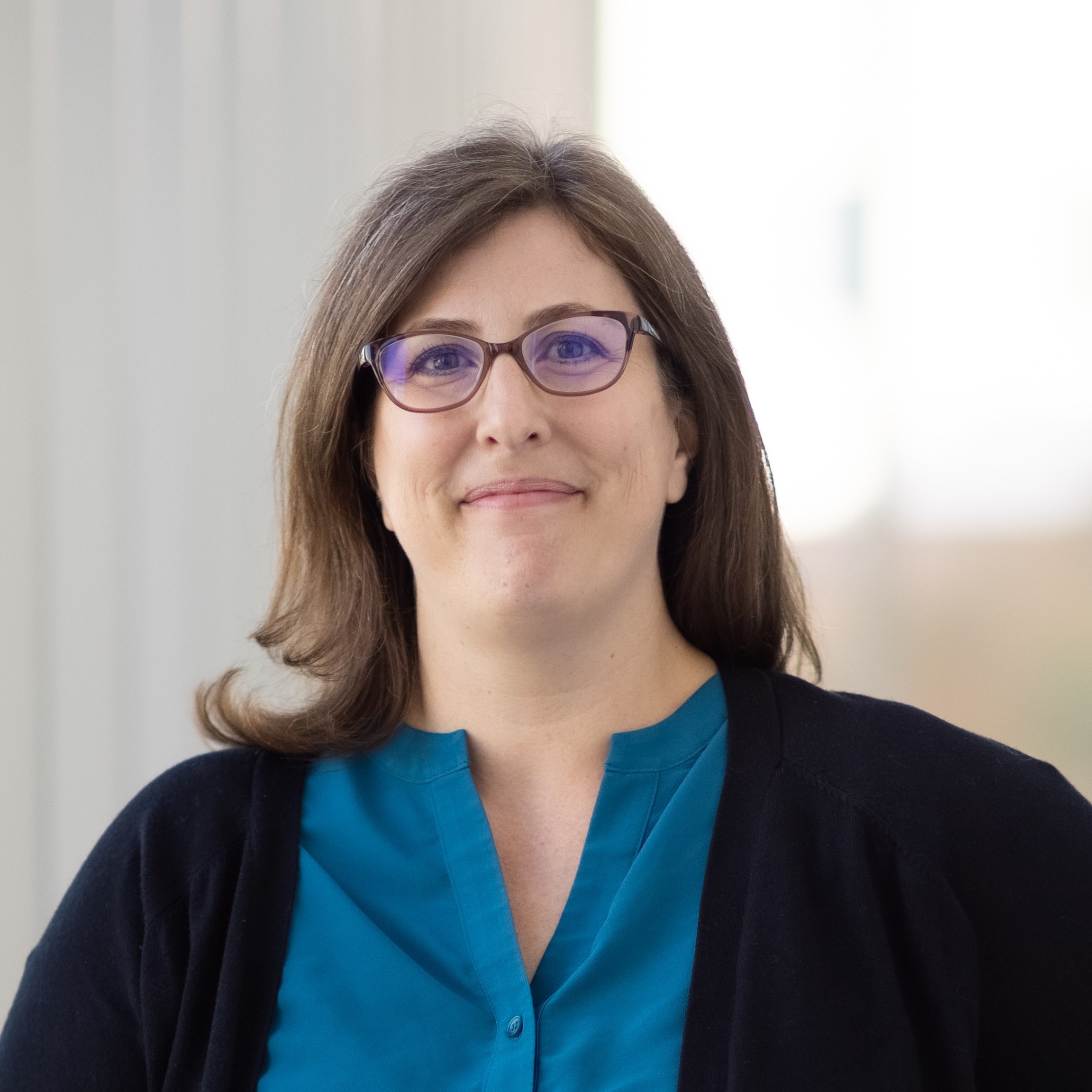 Portrait of Robin Whitney, a pale-skinned woman wearing a dark blue blouse and a black sweater. The background is out of focus and cream-colored.