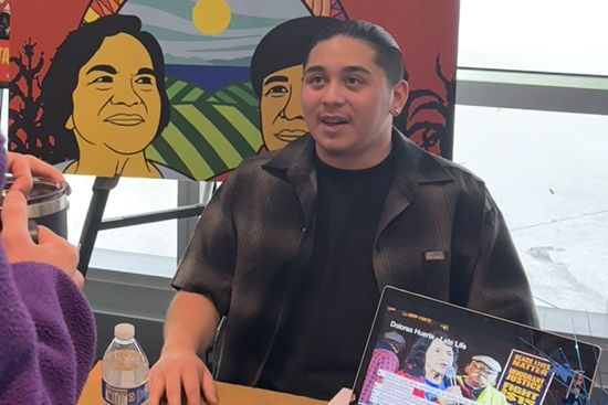 person presents at a table with computer edge in foreground and dolores huerta poster looking over shoulder