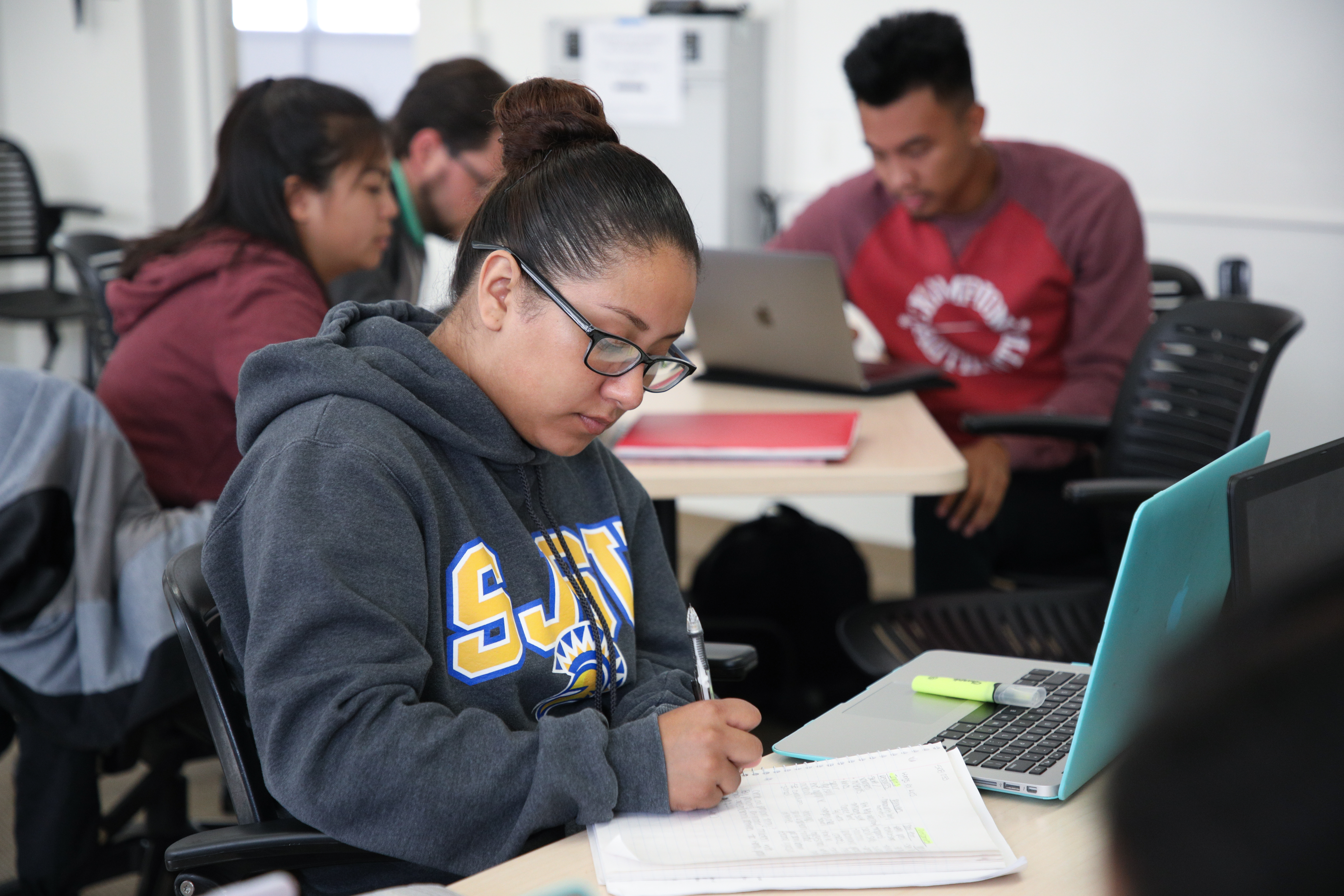 A female student wearing an SJSU sweatshirt sits and takes notes off a laptop screen. Other students are out of focus in the background doing the same.